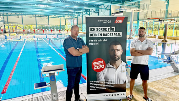 Zwei Personen stehen in einem Schwimmbad vor einem Roll-Up der Initiative